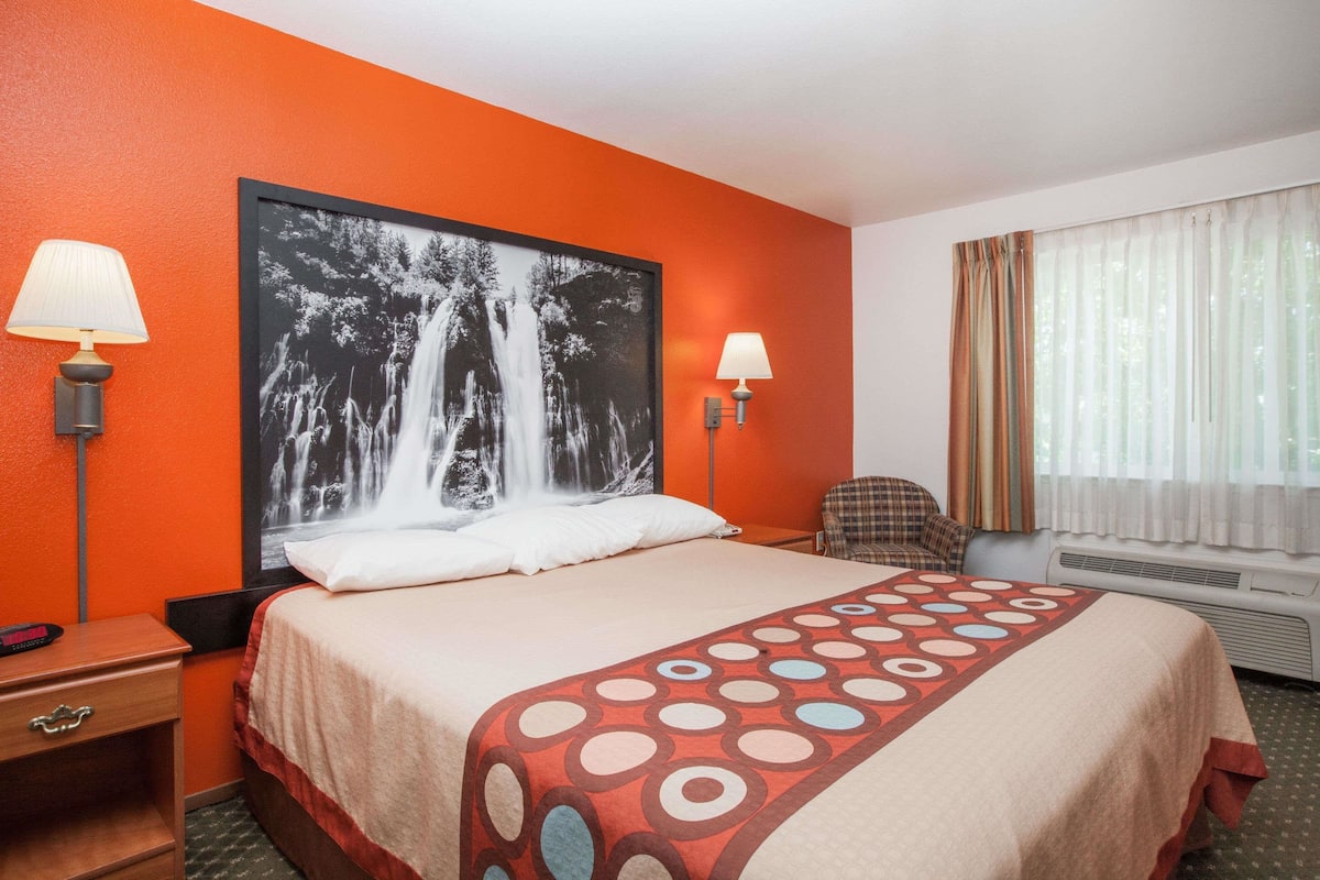 Camera d'albergo con un letto grande, due lampade sui comodini, una sedia, una finestra con tende e un condizionatore d'aria a parete.