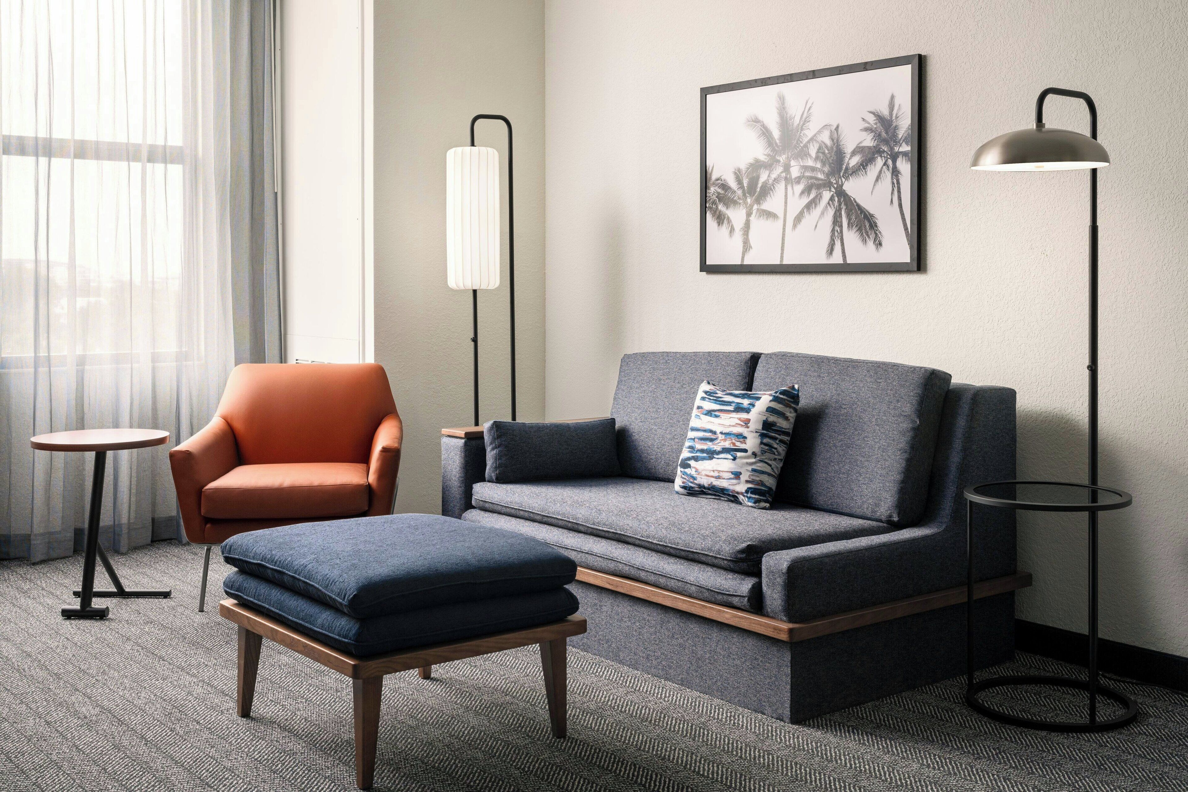 A modern hotel room with a grey sofa, orange armchair, and a small round table.