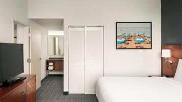 Habitación de hotel con cama, televisión sobre un aparador de madera, armario, baño y un cuadro con motivos playeros en la pared.