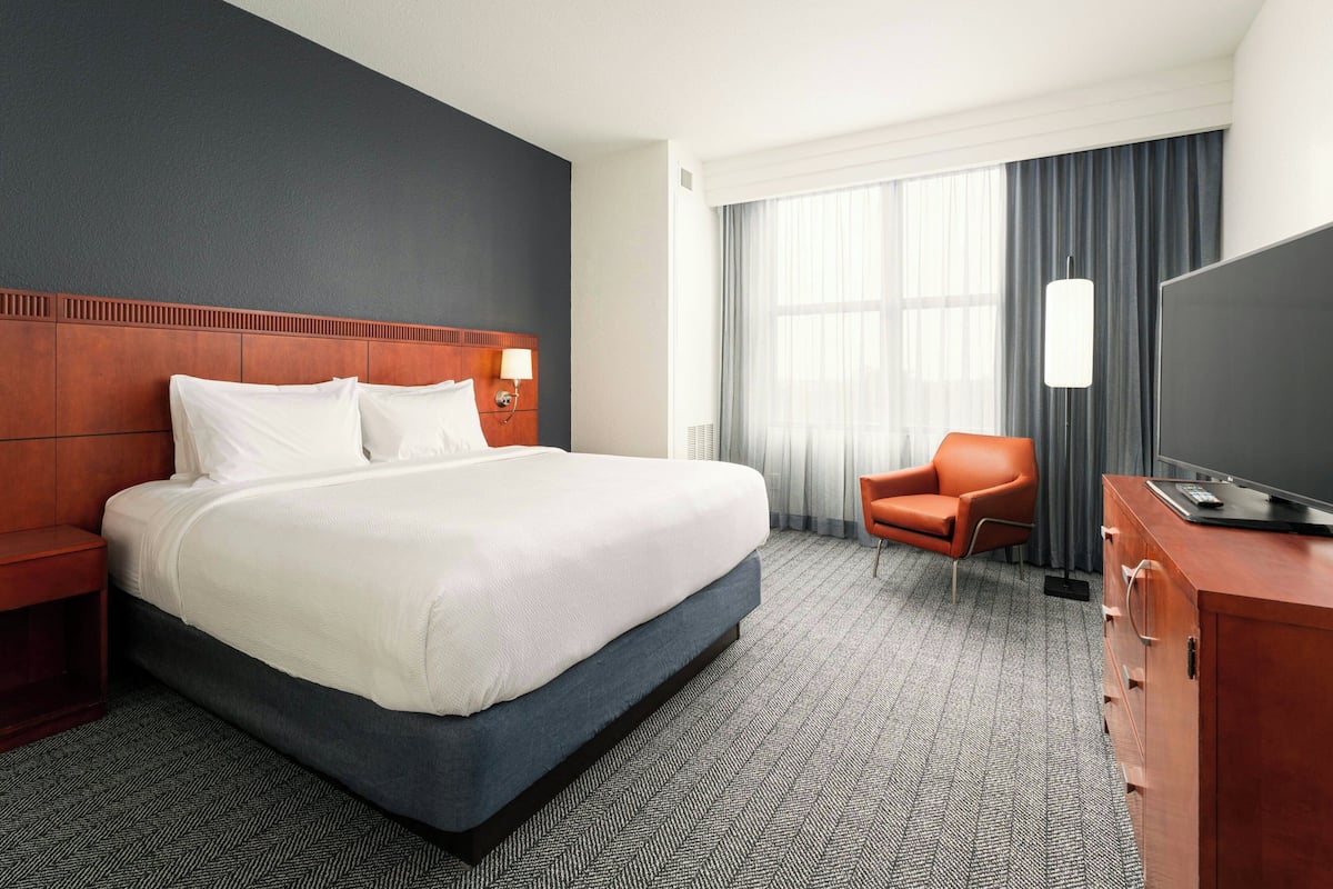 A hotel room with a large bed, a wooden headboard, a television on a wooden cabinet, a chair, and a large window with curtains.