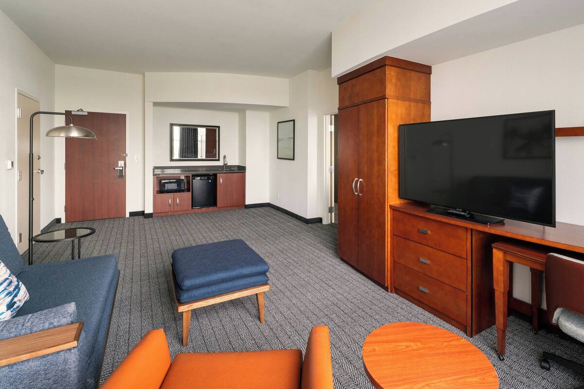 A hotel room with a sofa, ottoman, small table, TV, wooden cabinet, kitchenette, and a mirror.