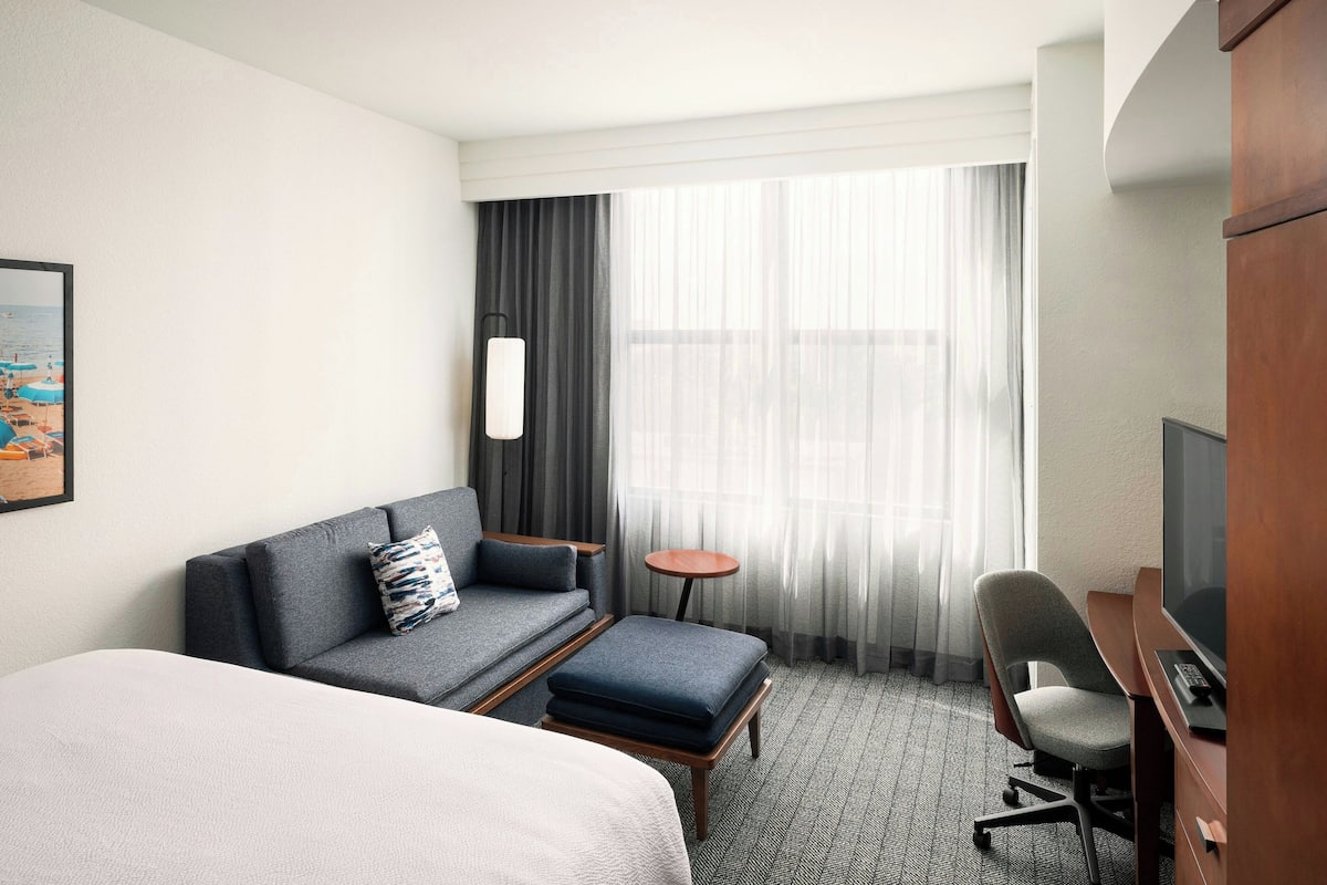 A hotel room with a bed, a sofa, an ottoman, a desk with a chair, a small table, and a large window with curtains.