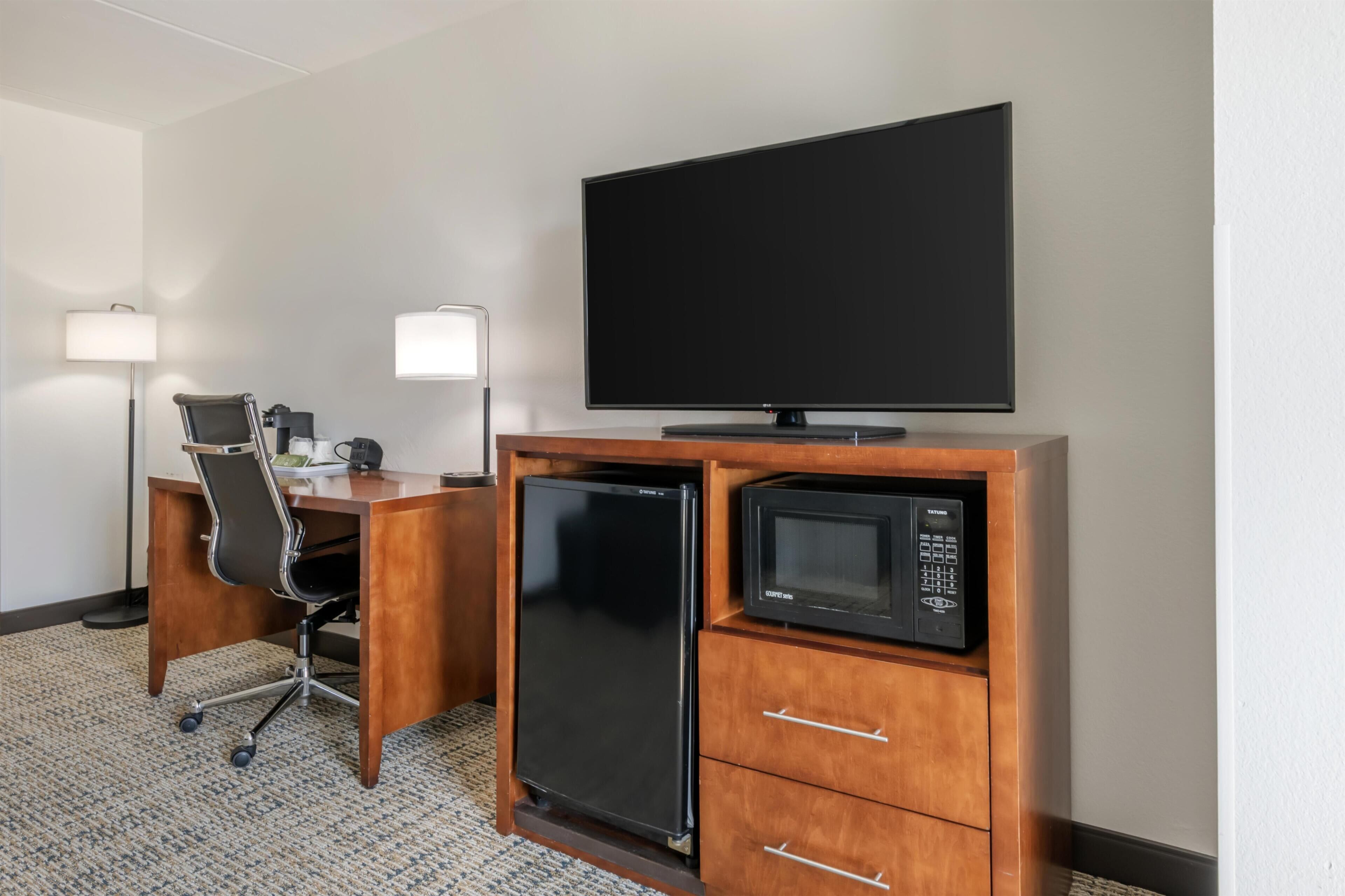 A hotel room with a desk, chair, flat-screen TV, mini-fridge, microwave, and a lamp.