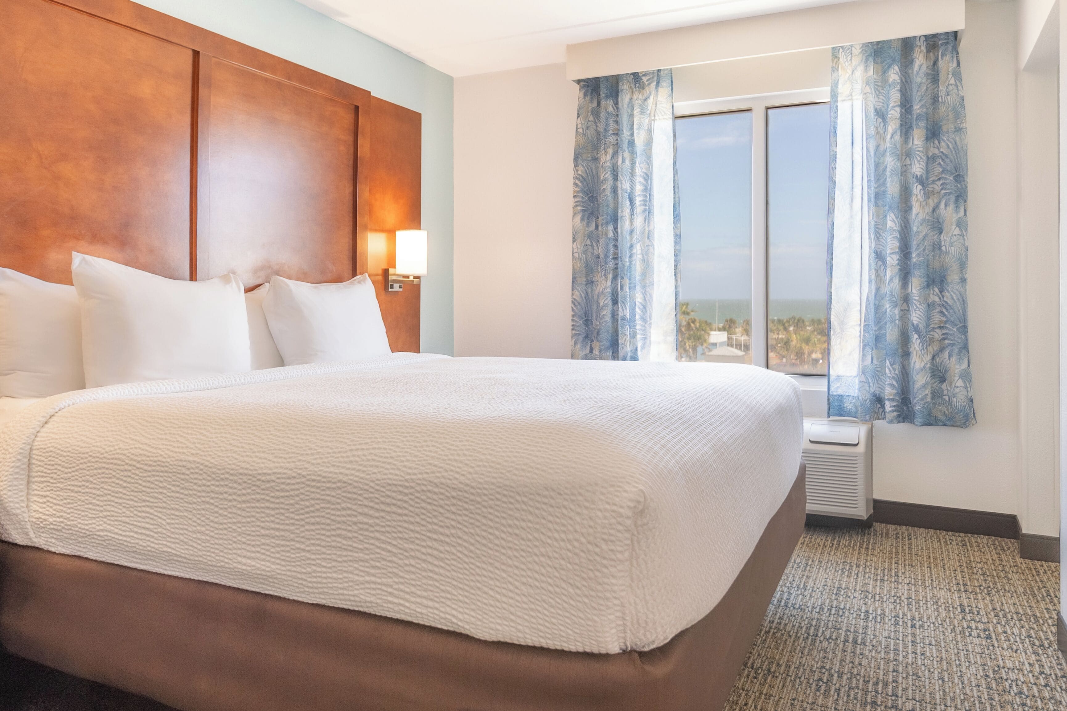 A hotel room with a large bed, a wooden headboard, a window with curtains, and a view of the city.