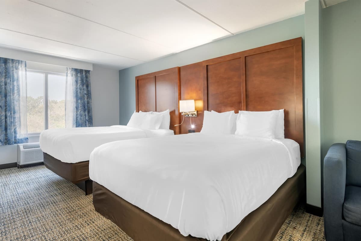 A hotel room with two beds, a nightstand with a lamp, a window with curtains, and a view of the outdoors.