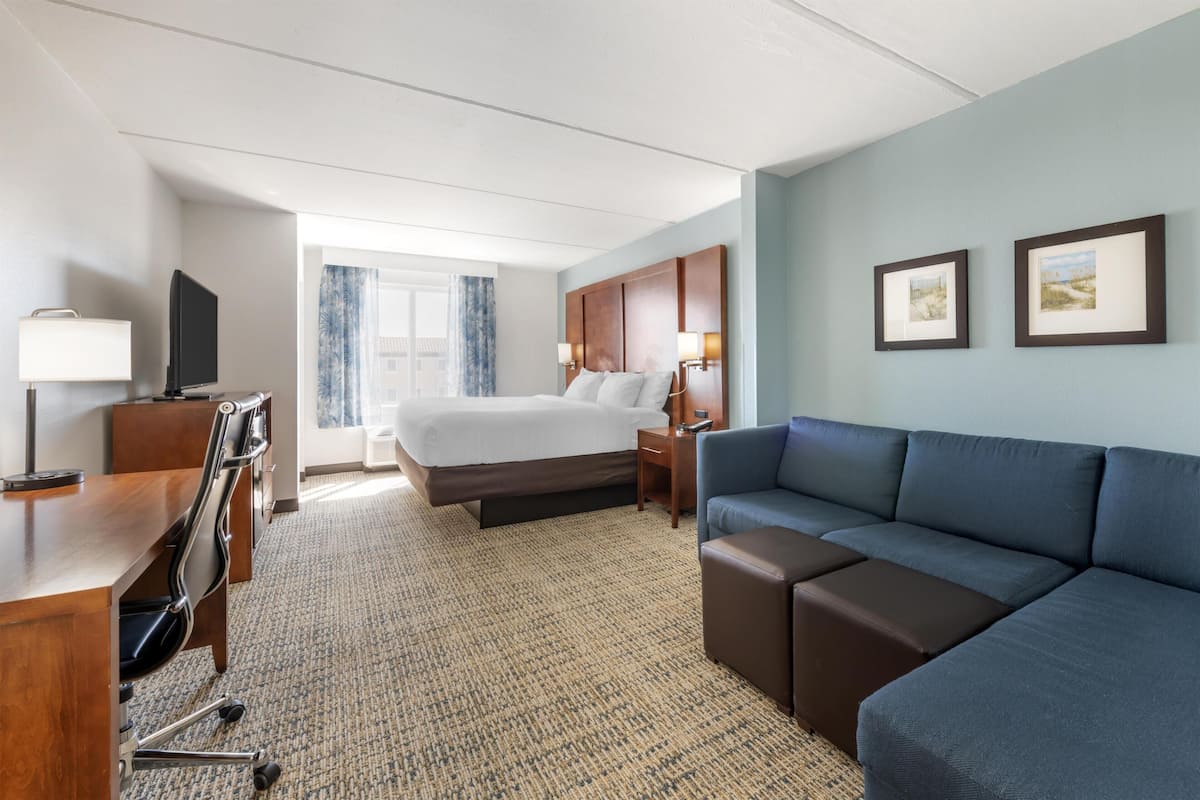 A hotel room with a bed, a sofa, a desk with a chair, a TV, and framed pictures on the wall.