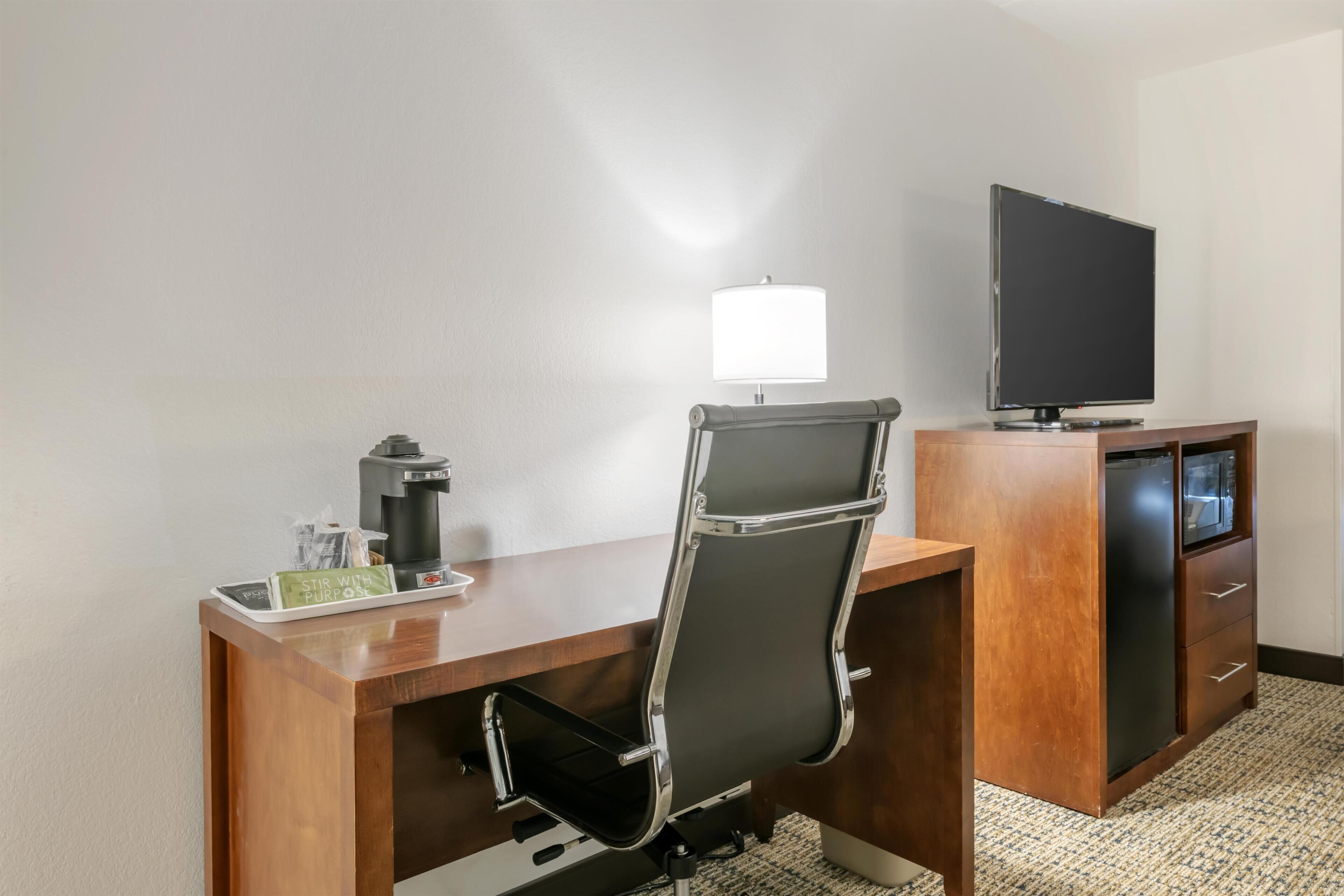A hotel room with a wooden desk, a black office chair, a flat-screen TV, a coffee maker, and a small tray with a coffee pot and a glass.