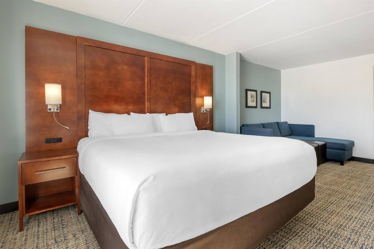 A hotel room with a large bed, a nightstand, a wall-mounted lamp, a blue sofa, and framed pictures on the wall.