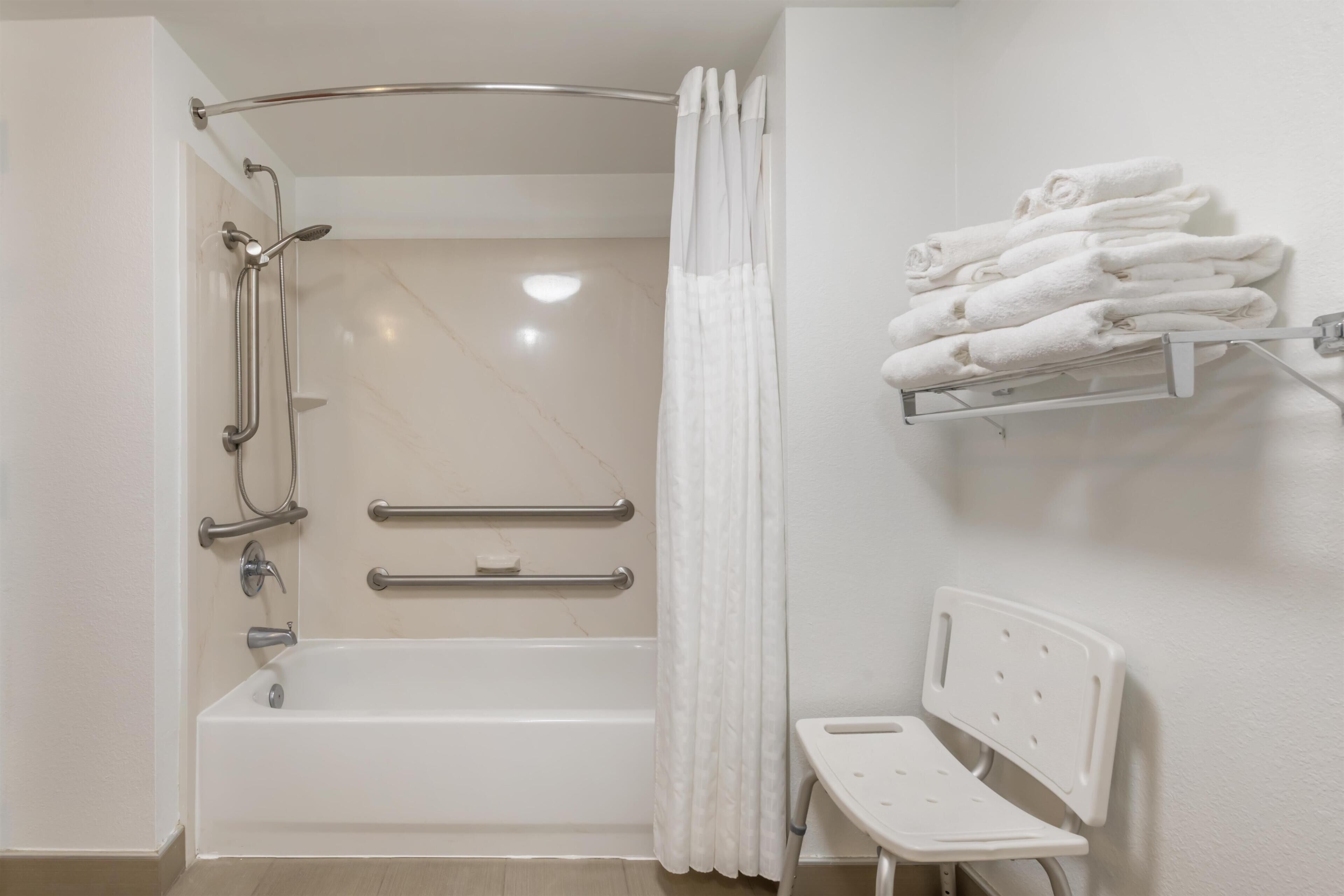 A bathroom with a bathtub, shower, handrails, a white bench, and a towel rack.