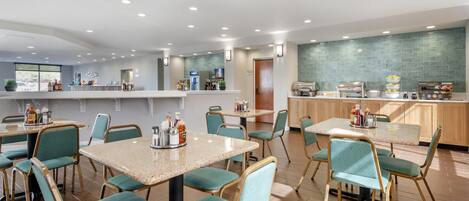 A modern hotel dining area with multiple tables and chairs, a kitchen counter, and a tiled backsplash.