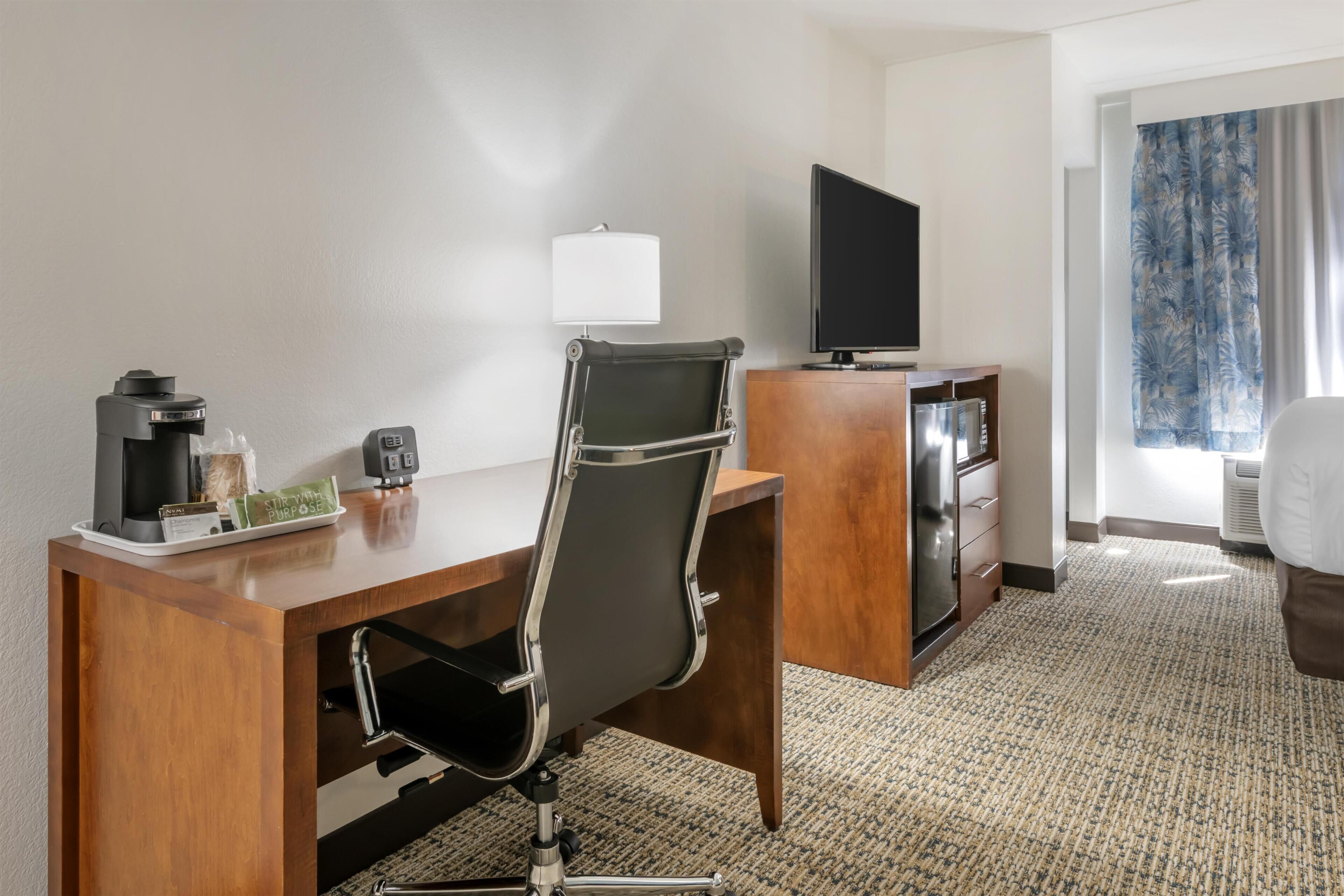 A hotel room with a wooden desk, a black office chair, a television, a coffee maker, and a window with curtains.