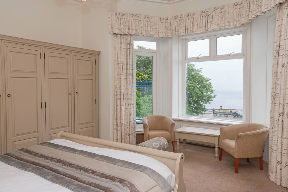 A bedroom with a bed, two chairs, a desk, and a large window with a view.