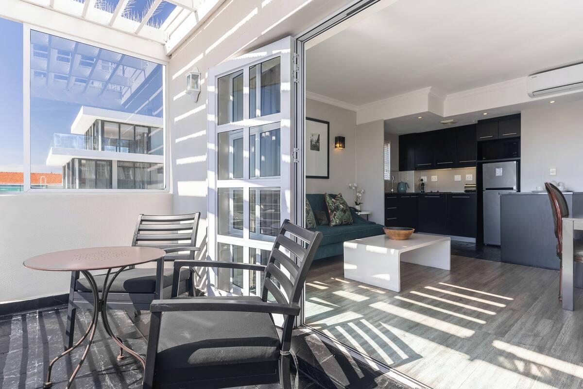 Un apartamento moderno con balcón, comedor con mesa y sillas, cocina con armarios oscuros y un ventanal que proporciona luz natural.