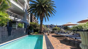 A swimming pool with lounge chairs, a palm tree, and a tiled walkway. - Romney Park Luxury Apartments (Cape Town)