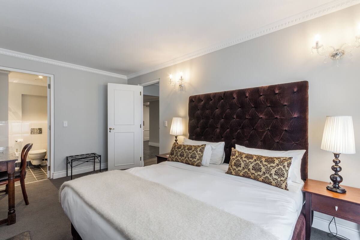 Habitación de hotel con una cama grande, una mesita de noche con lámpara, una silla y un escritorio con otra silla.