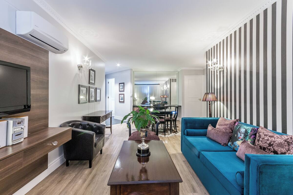 Una sala de estar moderna con un sofá azul, una mesa de centro, un televisor y un comedor al fondo.