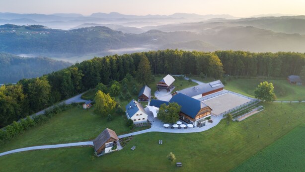 Une vue aérienne d’une propriété rurale comprenant plusieurs bâtiments, dont une grande grange et plusieurs constructions plus petites, le tout entouré d’une végétation luxuriante et de collines ondulantes.