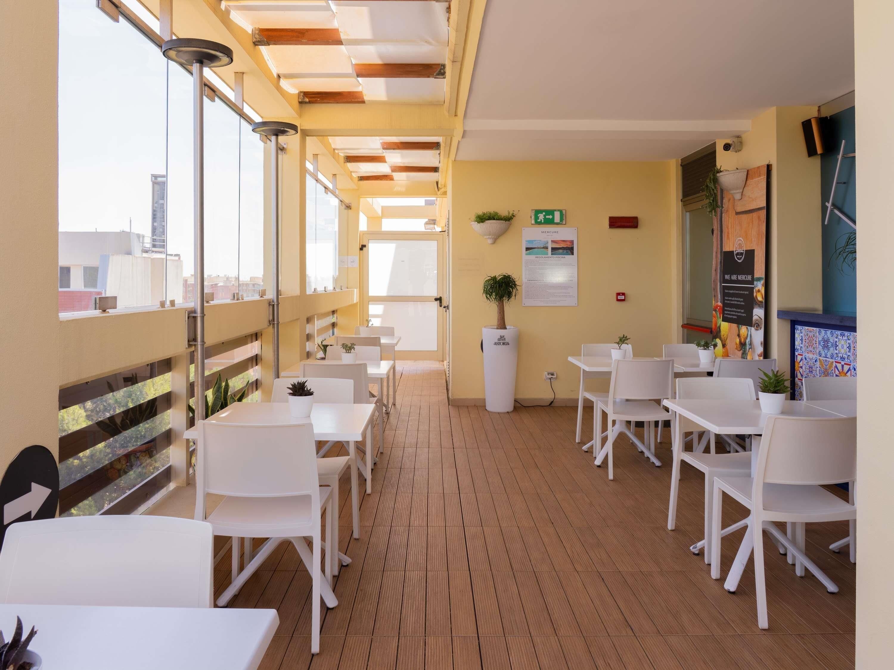 Un espacio comedor al aire libre con luz solar, mesas y sillas blancas, suelo de madera y vista panorámica de la ciudad.