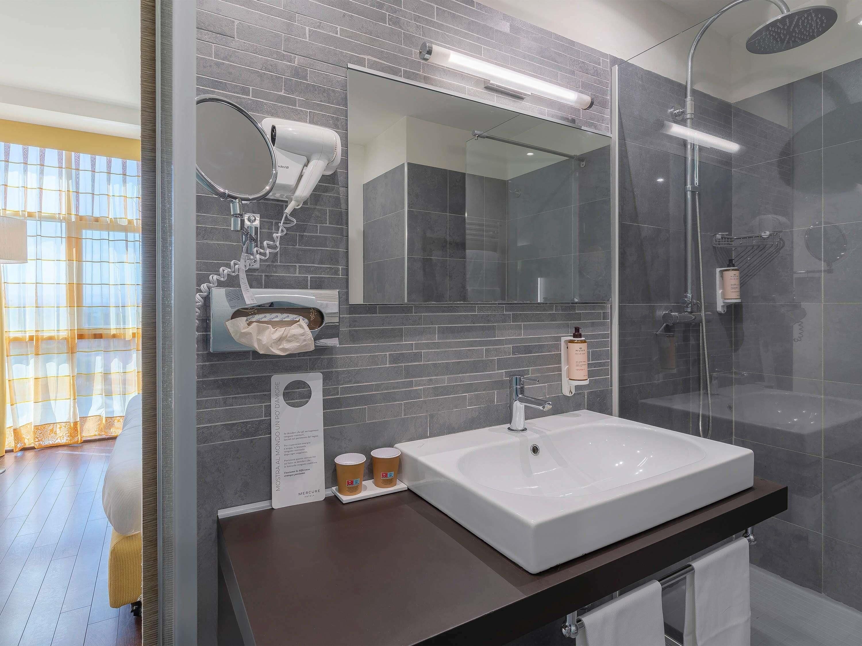 Une salle de bain moderne dotée d’un grand miroir, d’un sèche-cheveux, d’une douche en verre et d’un lavabo avec un porte-serviettes.