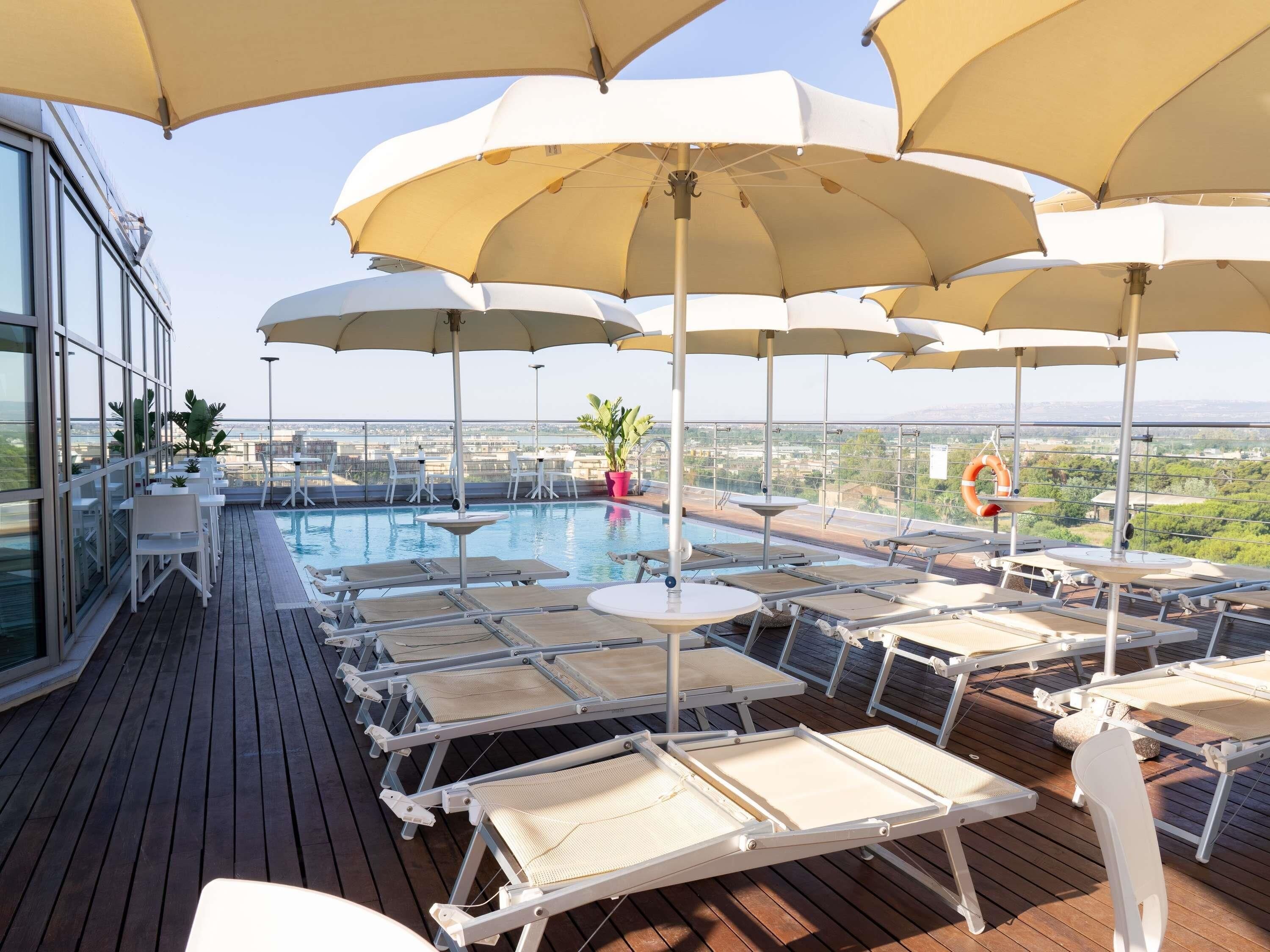 A rooftop pool area with sun umbrellas, lounge chairs, and a view of the cityscape.