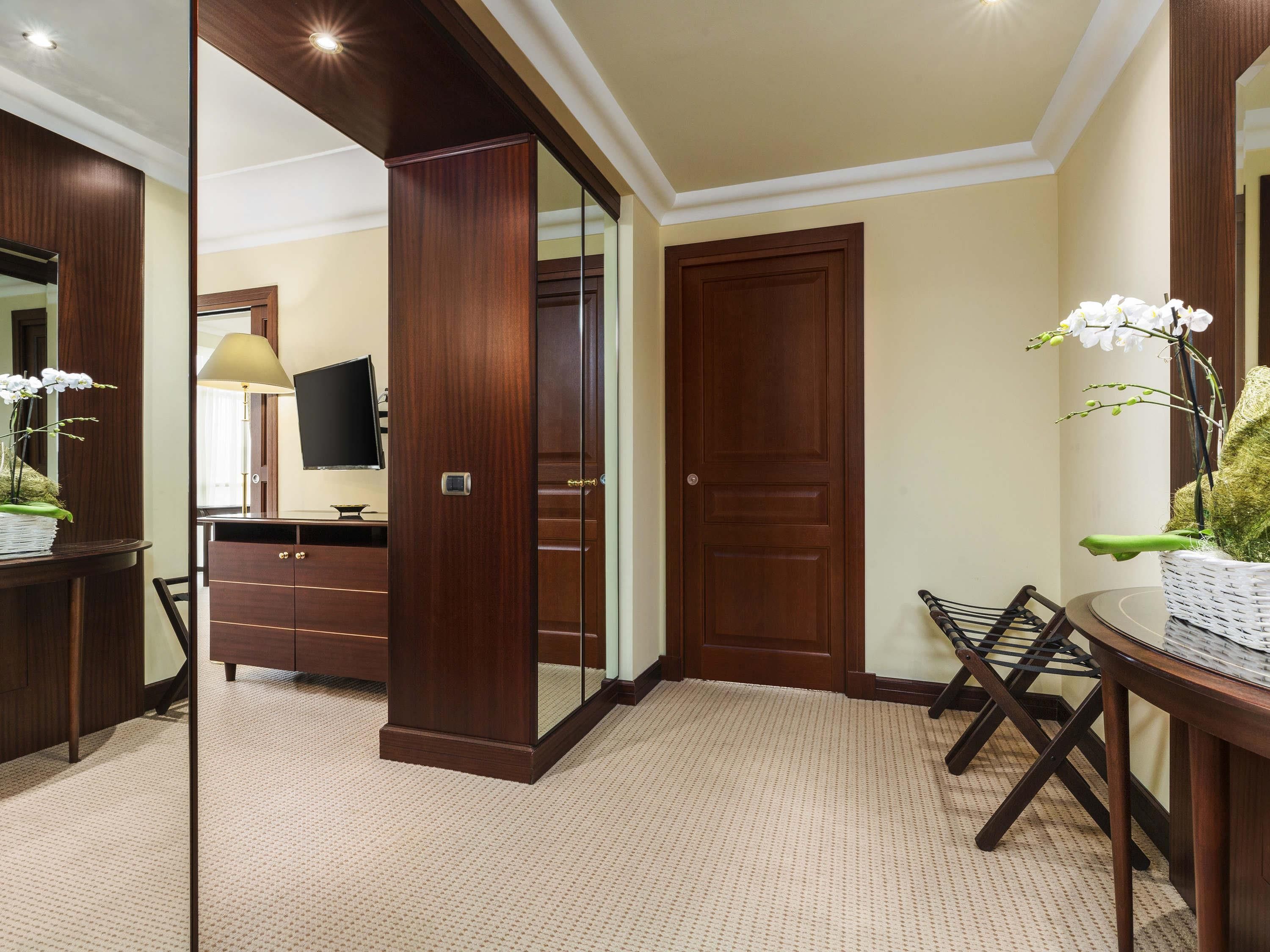A hotel room with a large mirror, a wooden wardrobe, a television, and a small table with a chair.