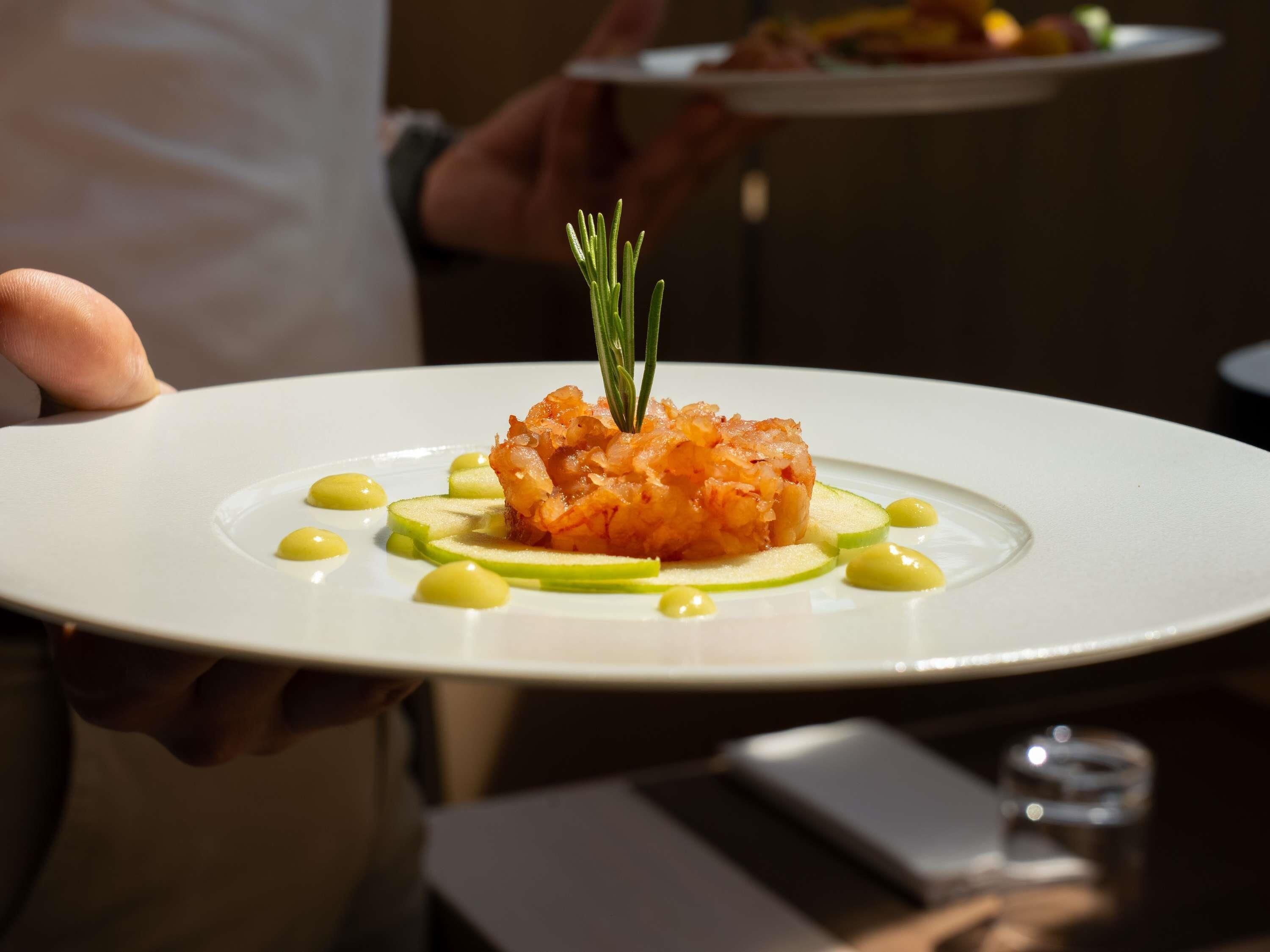 A person holding a white plate with a dish of food, garnished with a sprig of rosemary.