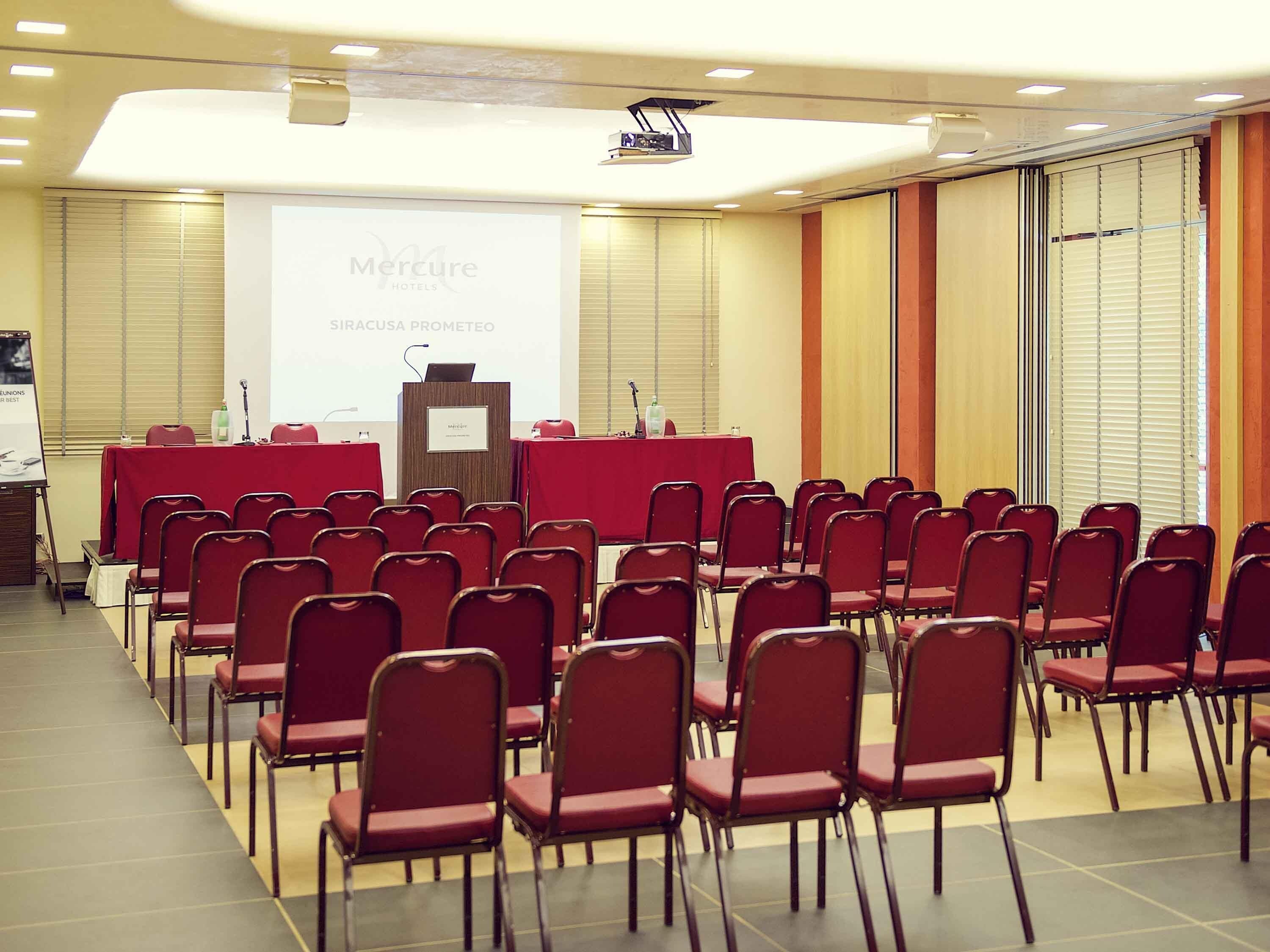 Un salón de conferencias con hileras de sillas rojas, un atril y una pantalla para presentaciones en Mercure Hotels.