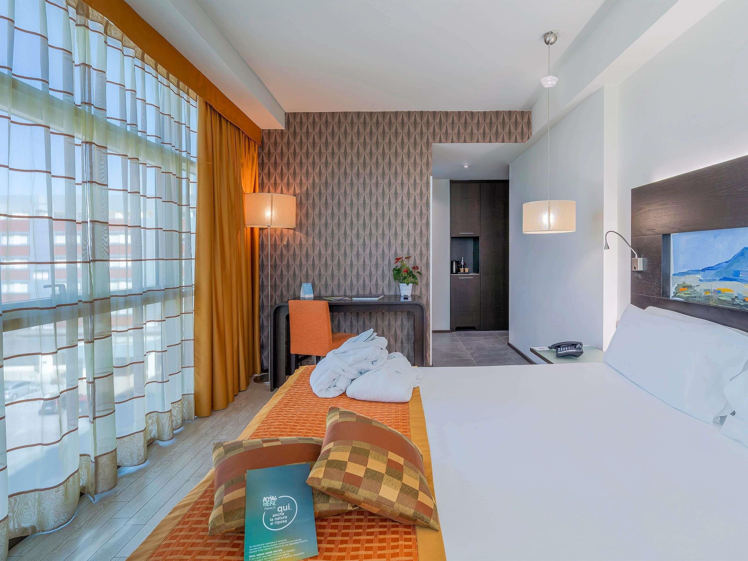 Una camera d'albergo con un letto, una scrivania, una sedia, una lampada, un quadro e vista sull'esterno.