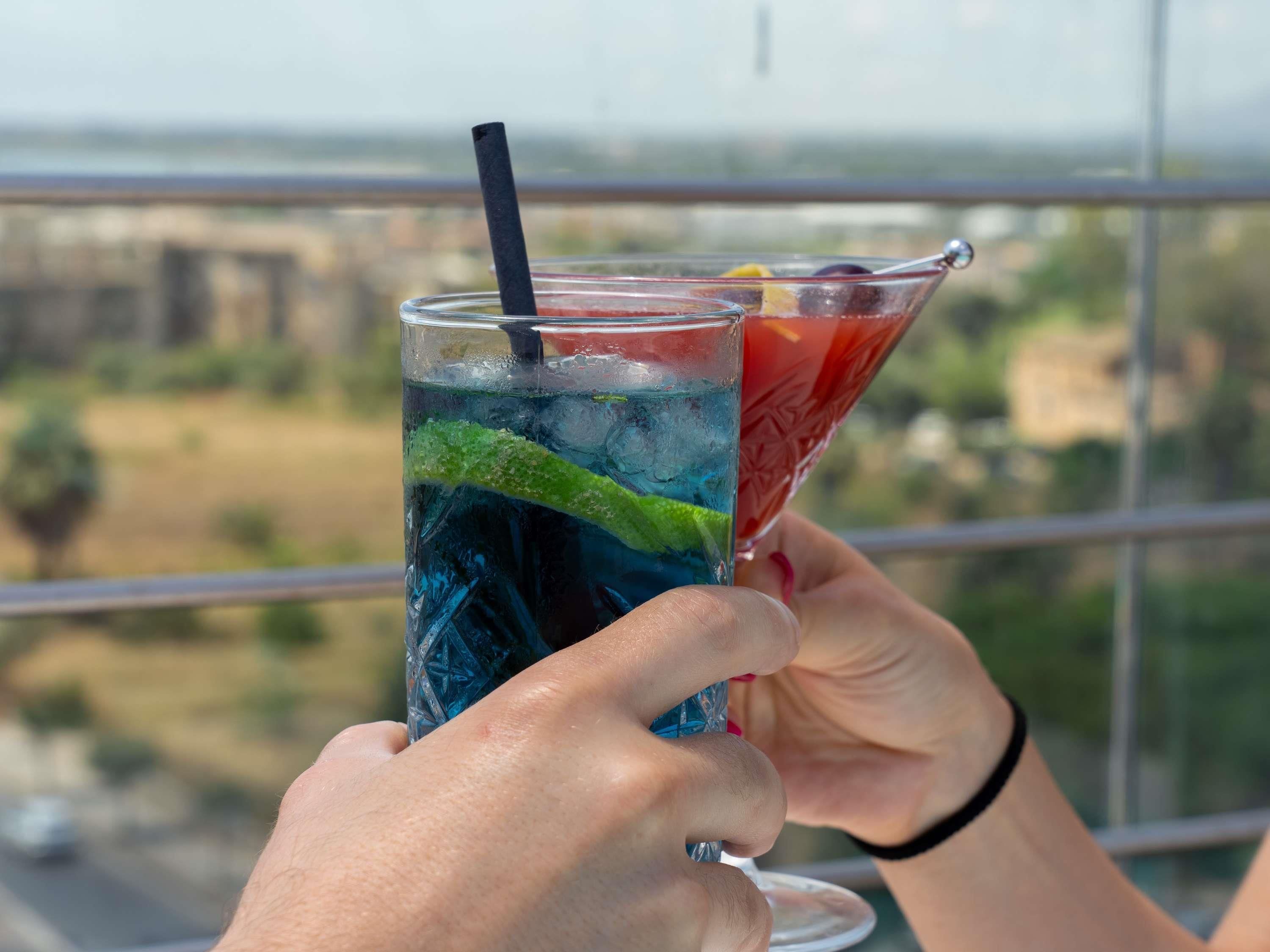 Two hands holding a tall glass and a martini glass with drinks, a black straw, and a slice of lime.