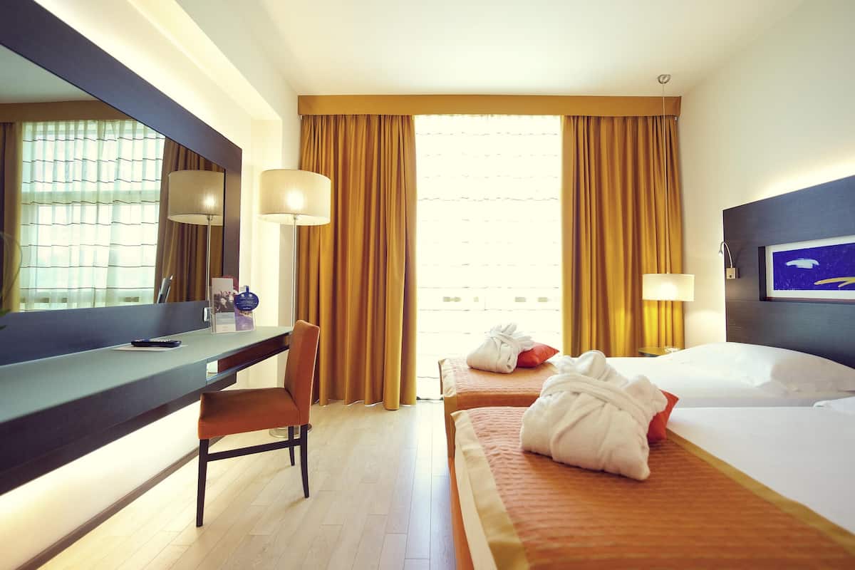 Ein Hotelzimmer mit Schreibtisch, zwei Betten, einem großen Fenster mit Vorhängen und einem gerahmten Bild an der Wand.