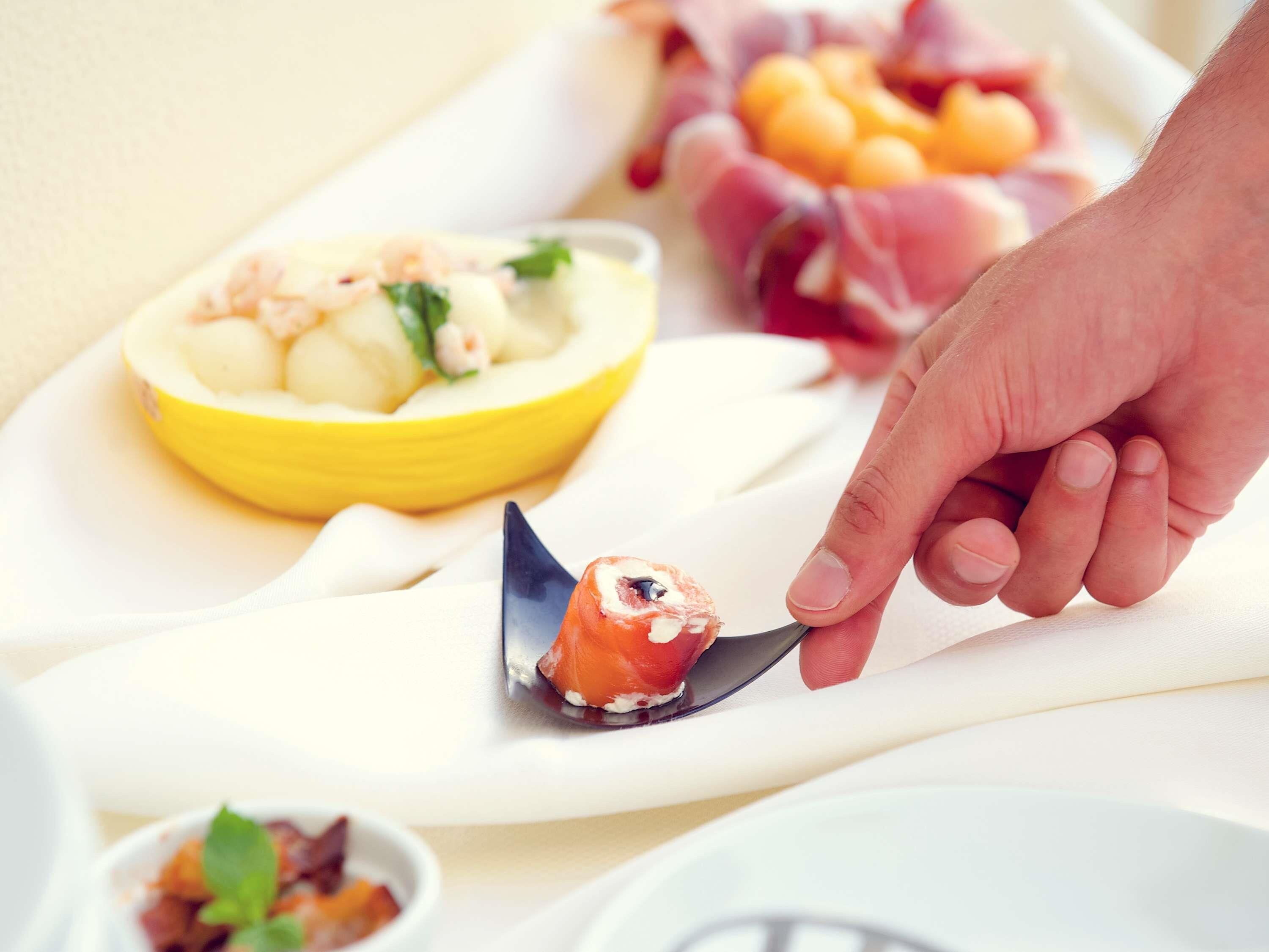 Una mano sosteniendo un trozo de sushi sobre un plato blanco, con un bol de fruta y un guiso de carne al fondo.