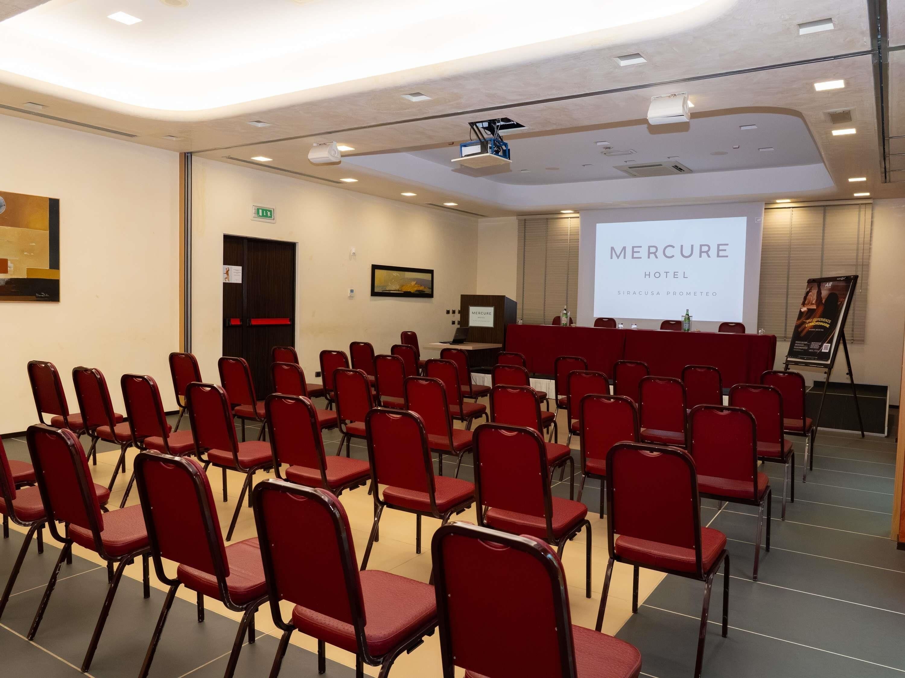 Un salón de conferencias con filas de sillas rojas, una pantalla de proyección y un atril.