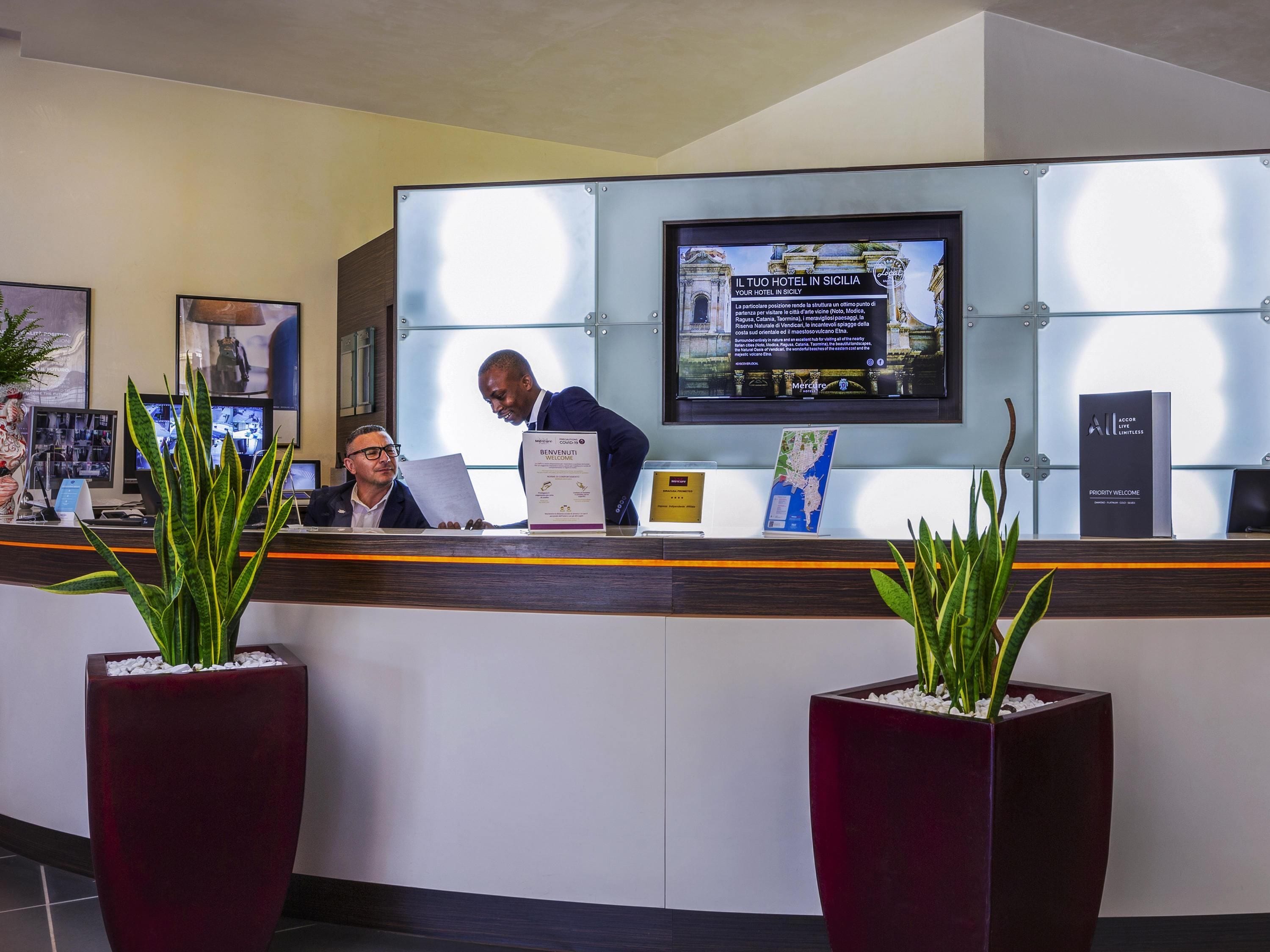 A hotel reception area with a reception desk, a large screen displaying information, and two potted plants.