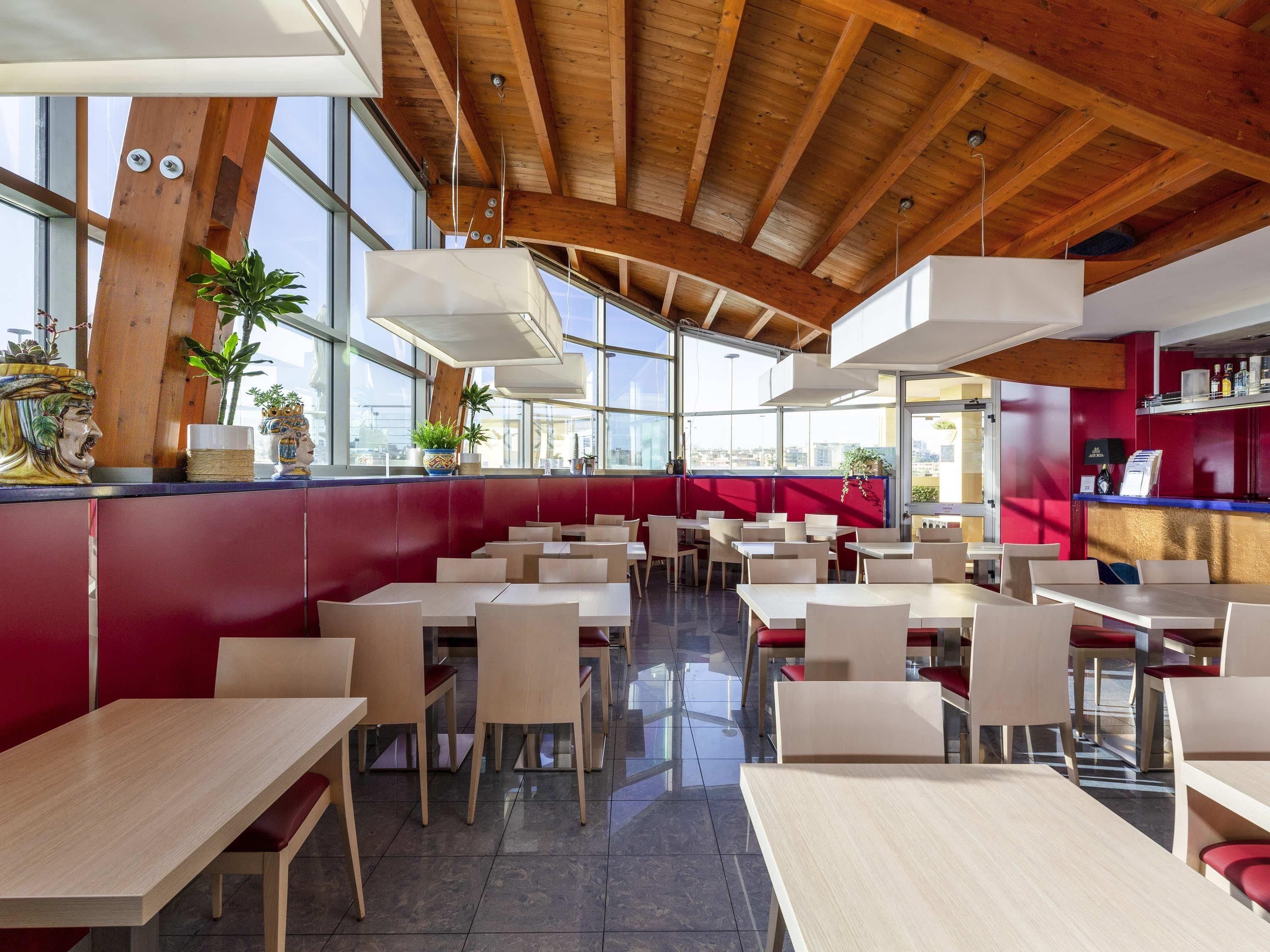 A modern restaurant with high ceilings, wooden beams, and large windows. Tables are arranged in a spacious layout with red accents on the walls.