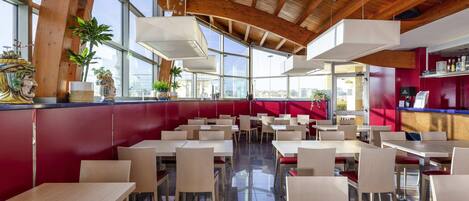 A modern restaurant with high ceilings, wooden beams, and large windows. Tables are arranged in a spacious layout with red accents on the walls.