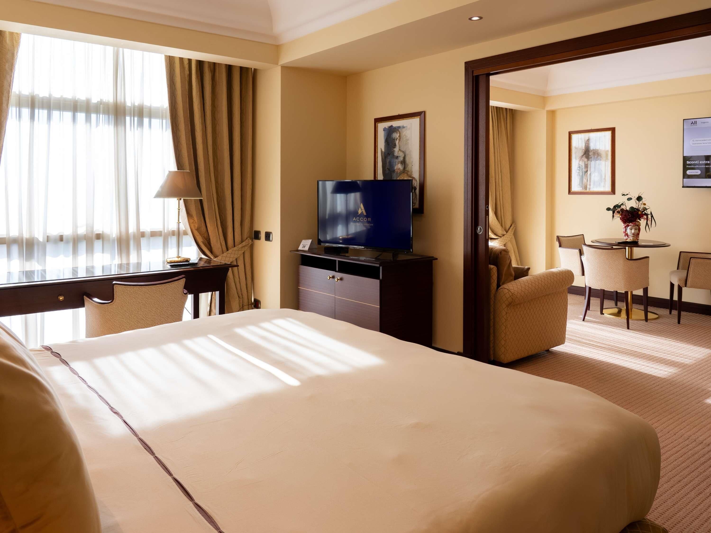A hotel room with a large bed, a desk with a television, a dining area with chairs, and a painting on the wall.