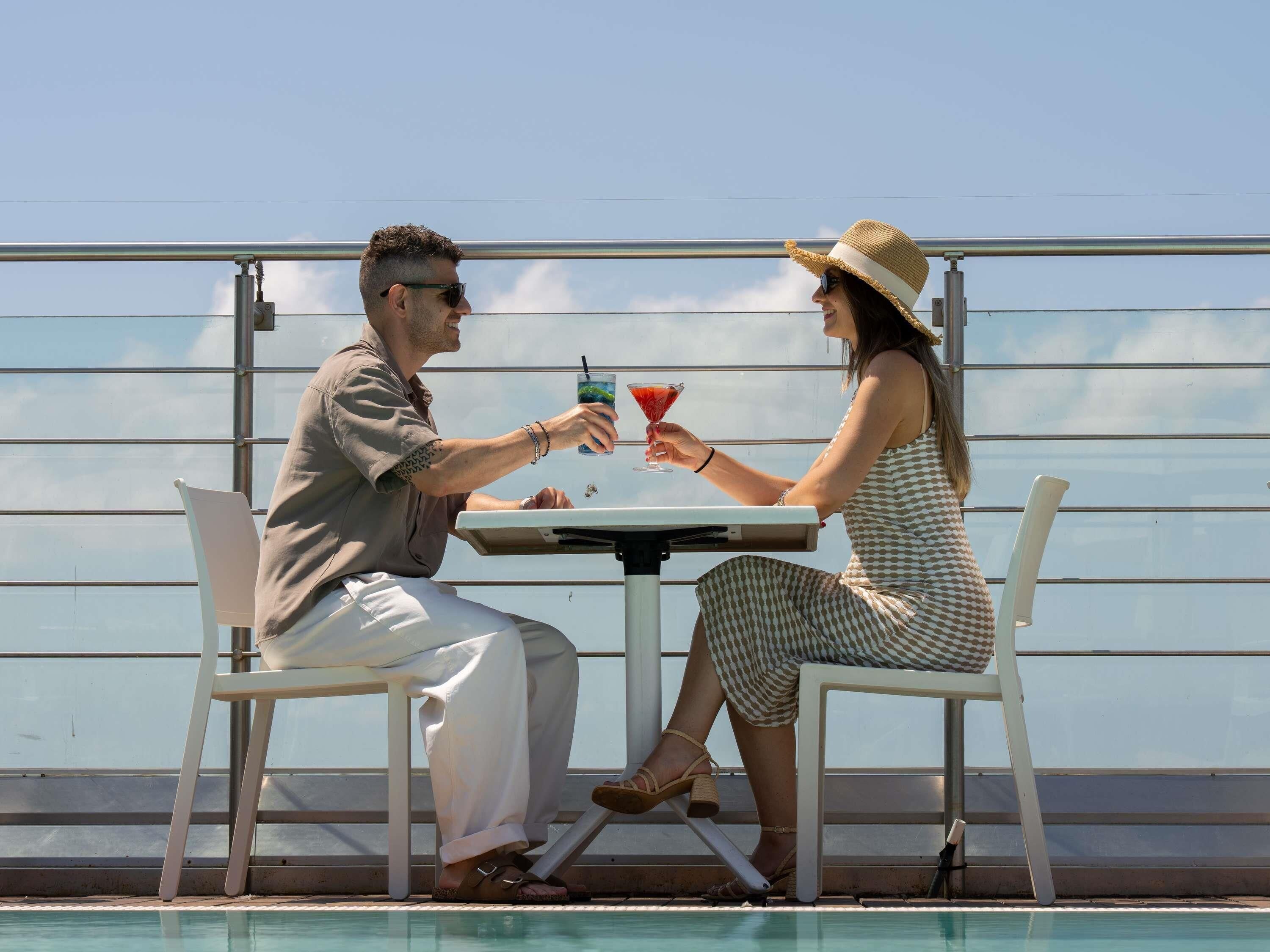 Two people seated at a table with a drink, overlooking a pool.