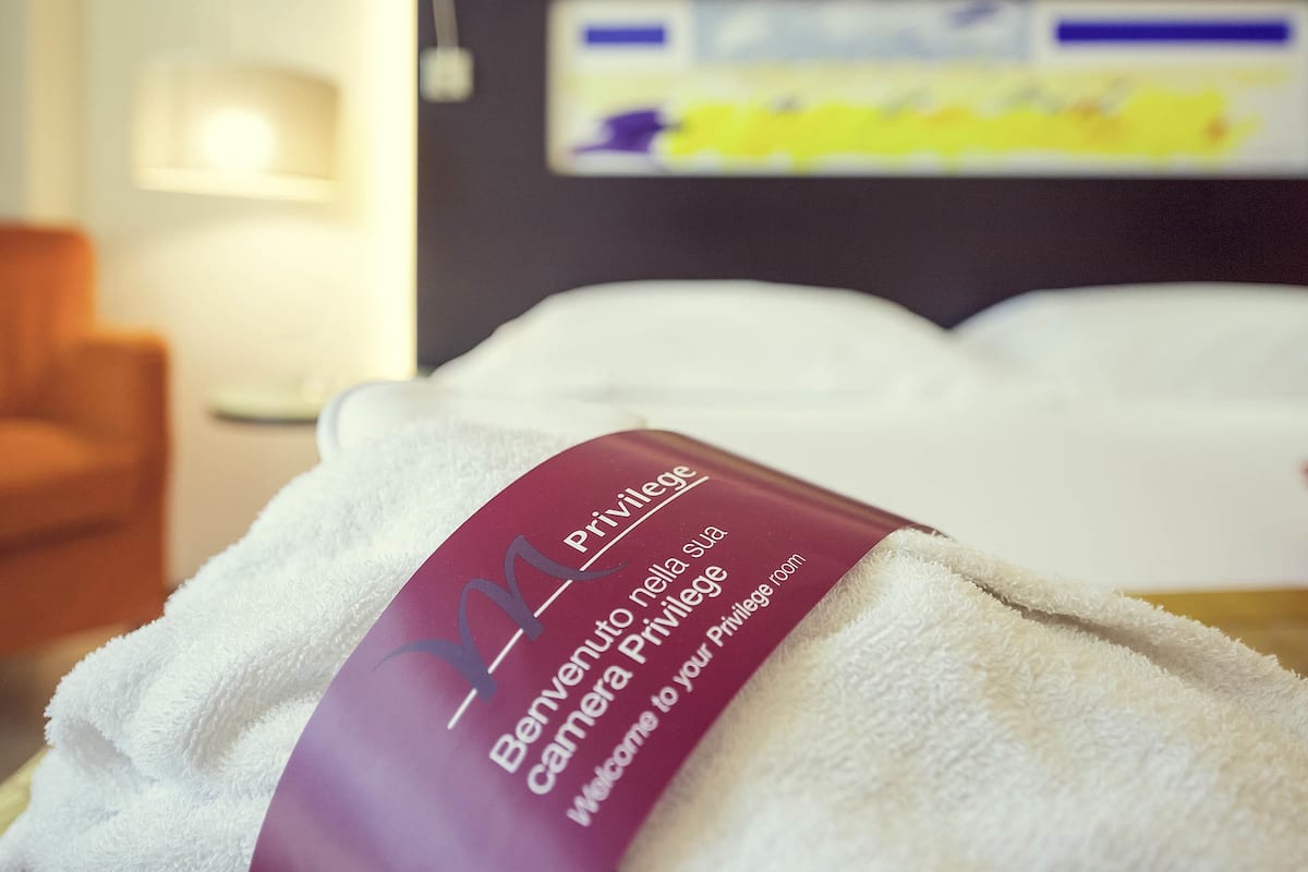 Ein Hotelzimmer mit einem ordentlich gefalteten weißen Handtuch, einem violetten Etikett mit der Aufschrift 'Benevento Privilegia' und einem Bett mit weißer Bettwäsche.