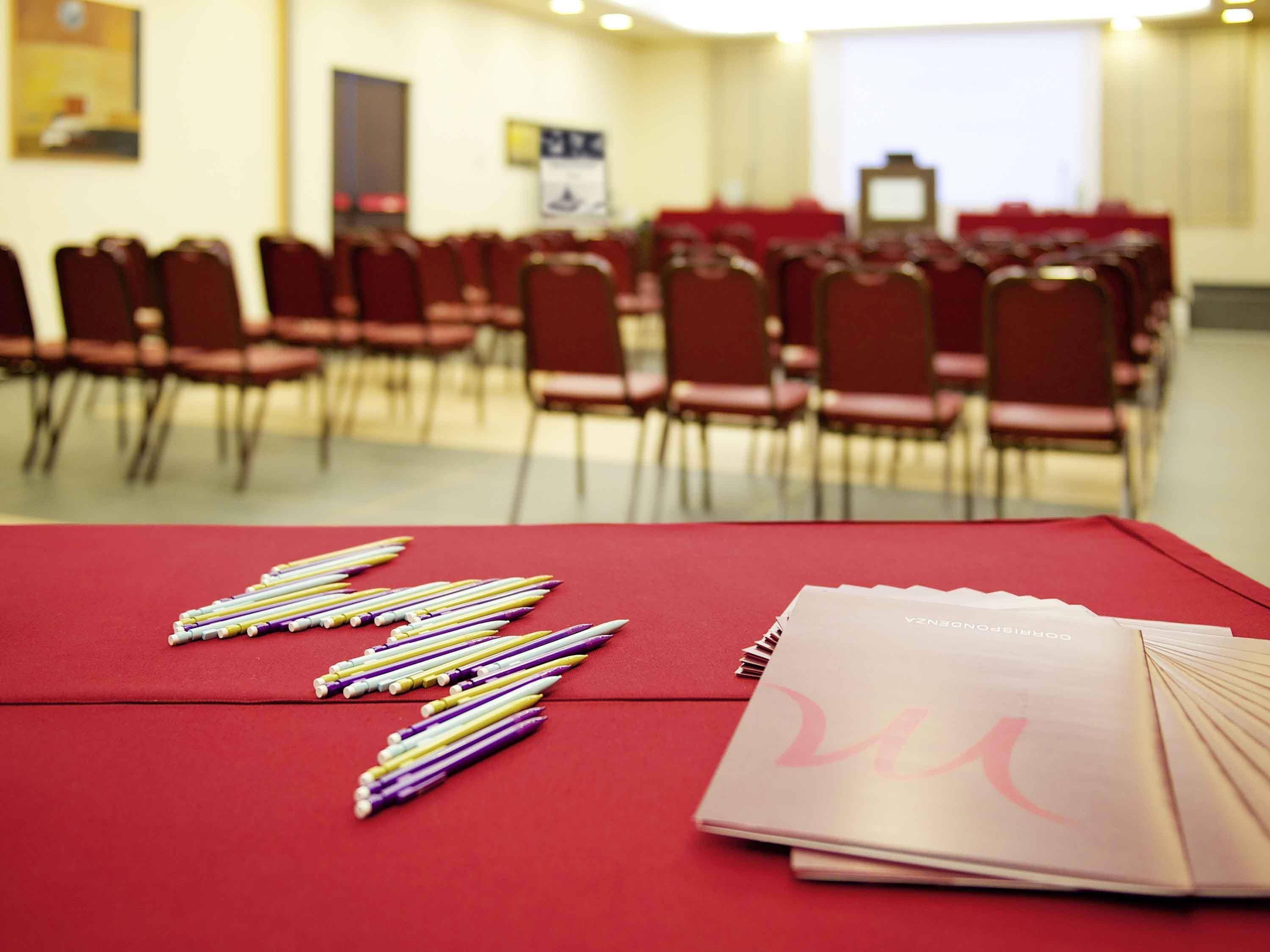 Una sala conferenze con file di sedie rosse, un tavolo con strumenti di scrittura e una cartella, e uno schermo per proiettore sullo sfondo.