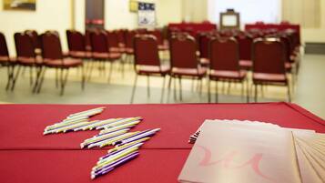 A conference room with rows of red chairs, a table with writing instruments and a folder, and a projector screen in the background.