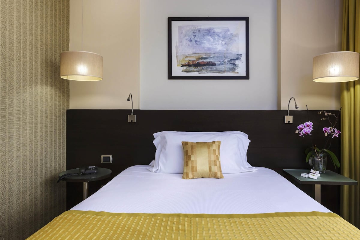 Ein Hotelzimmer mit einem Bett, zwei Nachttischlampen, einem Nachttisch, einem Telefon, einem Bild an der Wand und einer Blumenvase.