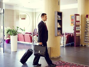 A man in a suit walking through a lobby with a briefcase, red seating, and a bookshelf. - Mercure Siracusa Prometeo (Syracuse)