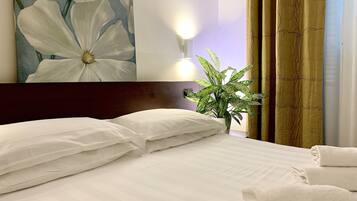 Een hotelkamer met een bed, kussens, een bloemschilderij, een wandlamp, een plant en gordijnen.
