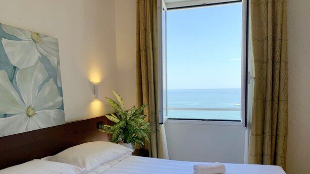 A hotel room with a bed, white pillows, a potted plant, and a view of the sea through a window. - Hotel Corallo (Imperia)