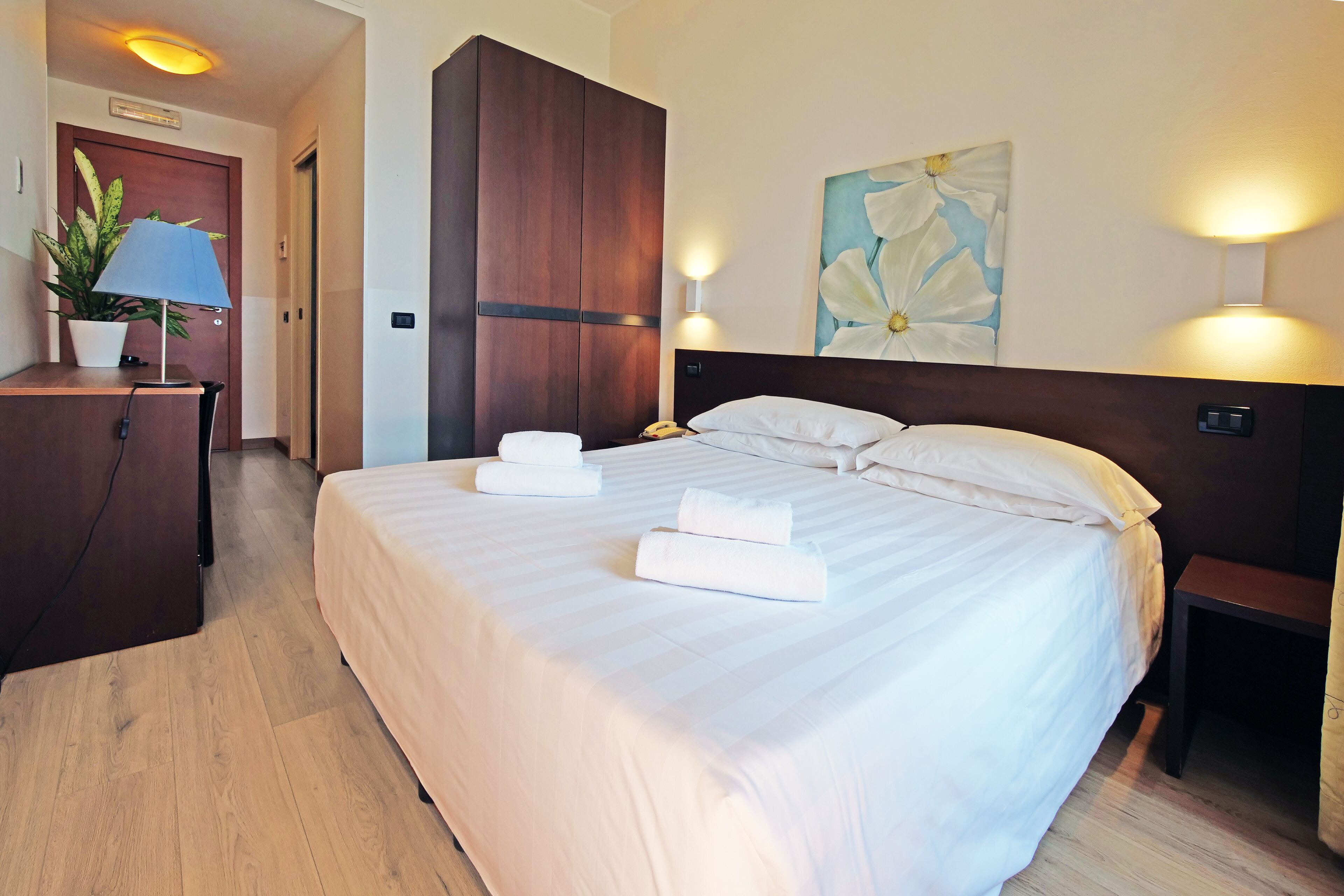 A hotel room with a large bed, a desk with a lamp, a potted plant, and a painting on the wall.