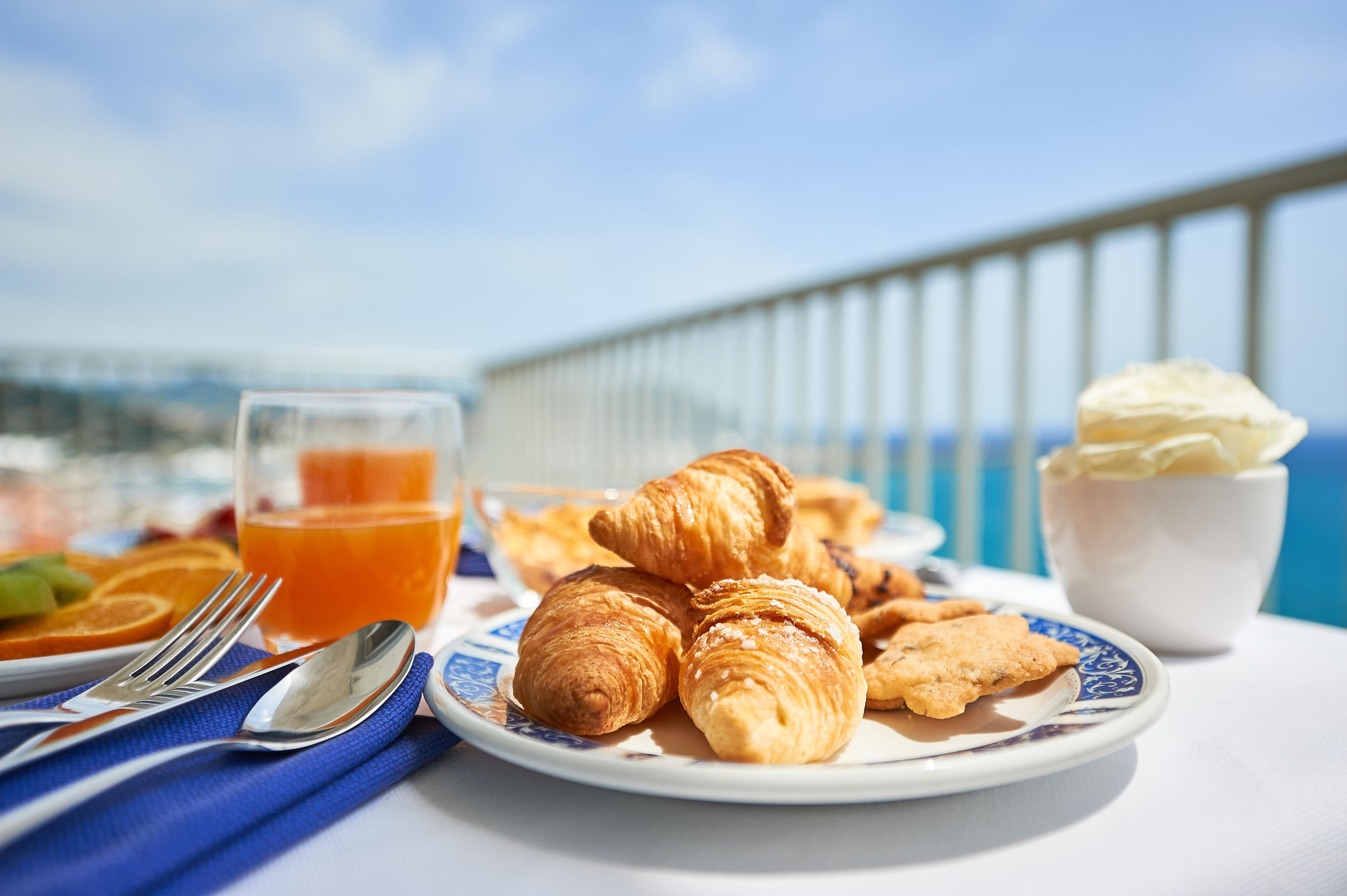 A breakfast setting with croissants, a glass of orange juice, and a bowl of ice cream on a table with a view of the sea.