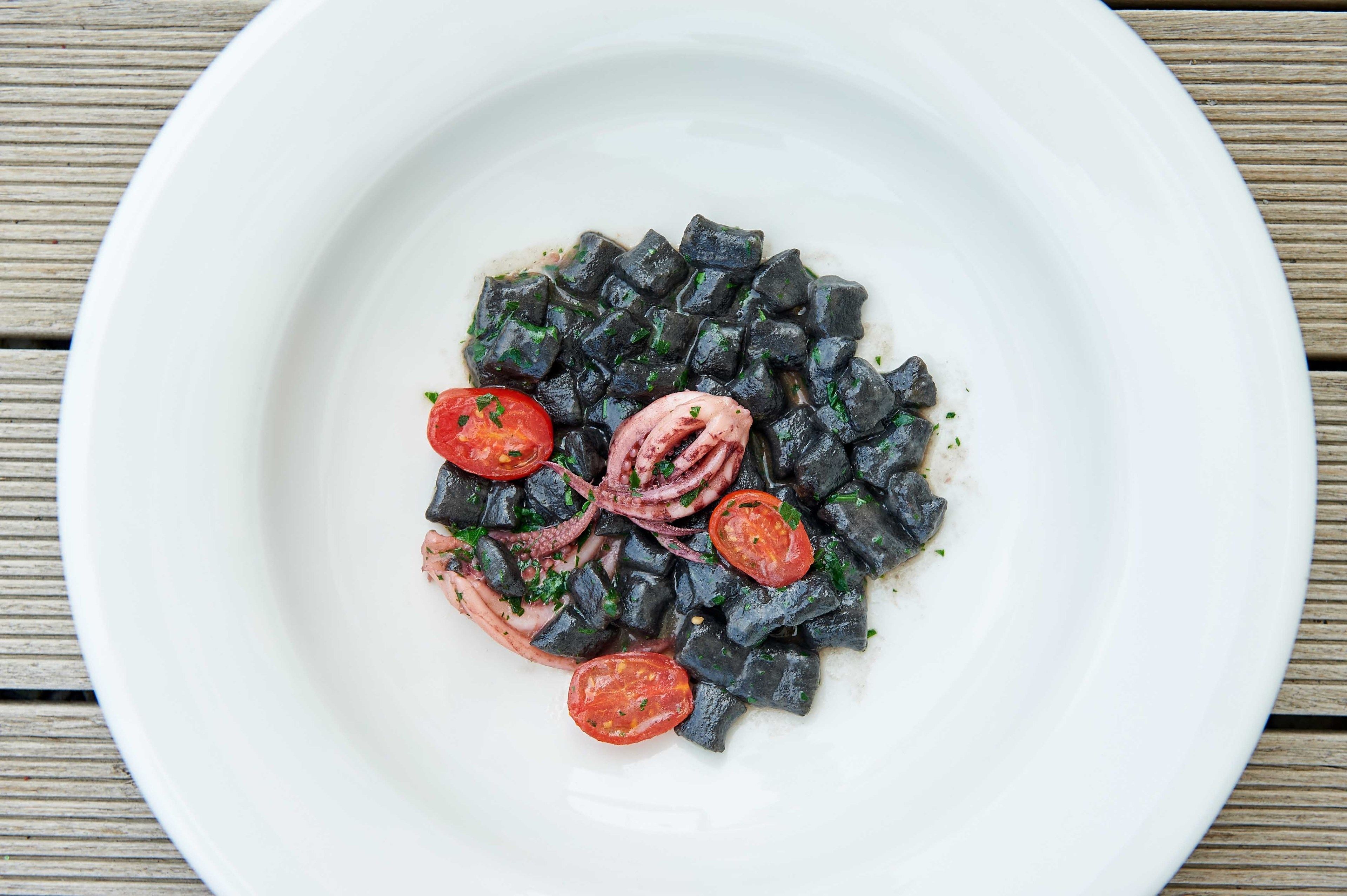 Plato blanco con pasta de tinta de calamar negro, tomates cherry y aros de calamar sobre una mesa de madera.