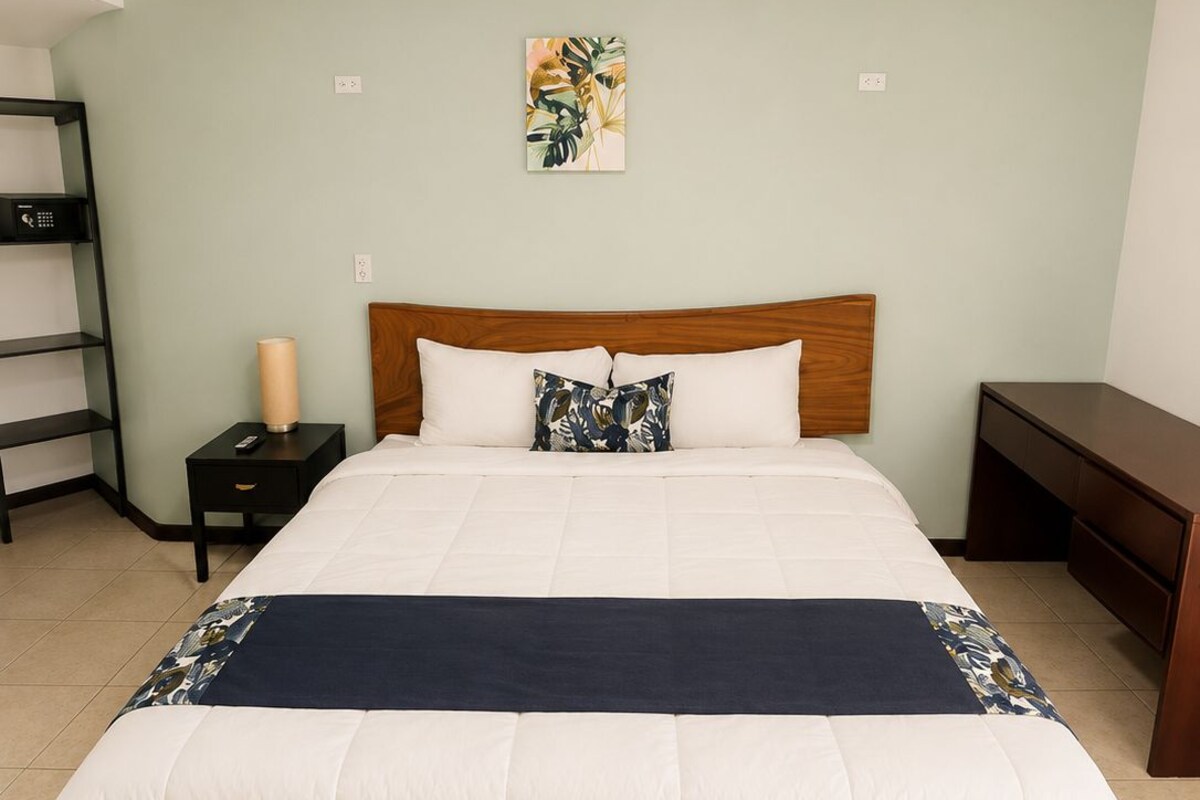 A hotel room with a wooden bed, a bedside table, a lamp, and a wall-mounted artwork.