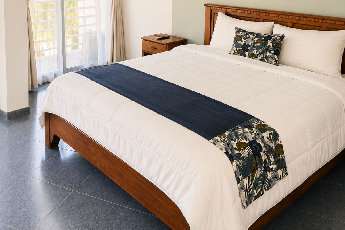 A bedroom with a large bed, a nightstand, a painting on the wall, and a view through a window with curtains.