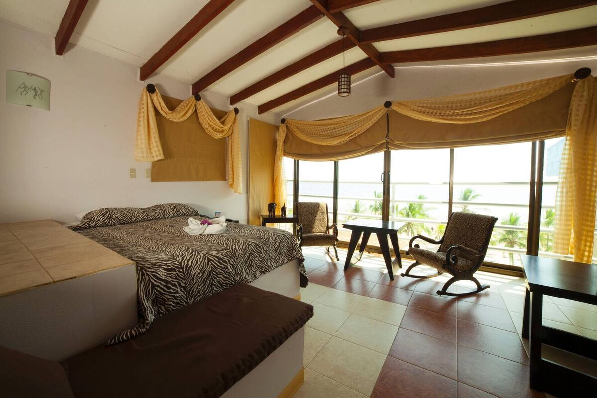 A room with a bed, two chairs, a table, and large windows with curtains.
